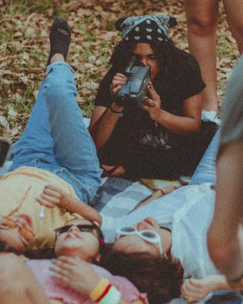 A group of young people are having a picnic in a park while one films them. 