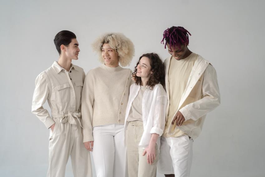 A diverse group of four young people smiling and conversing arm in arm, all wearing white against a light grey backdrop.