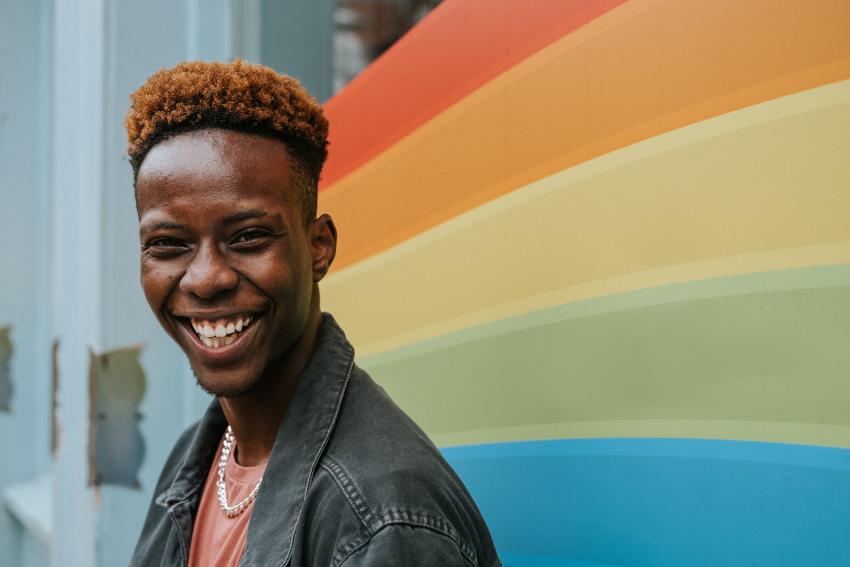 A young person smiling and laughing in front of a window displaying a Rainbow Pride flag.