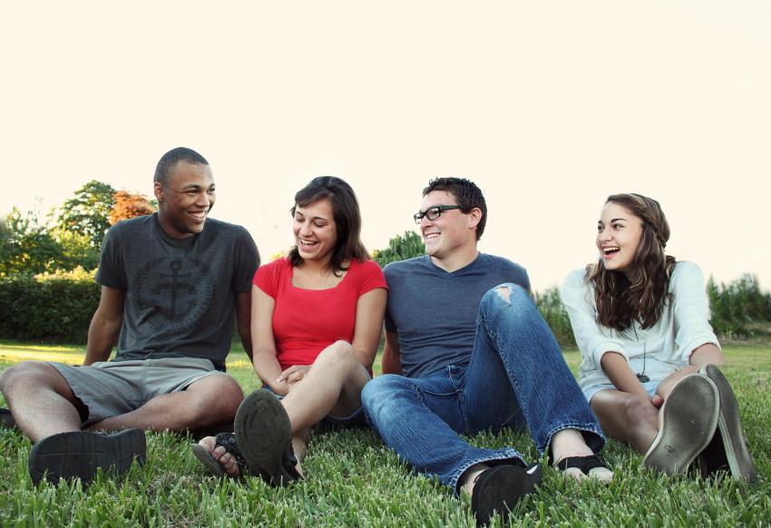 Four young people are sat in a row smiling and laughing in a park. 