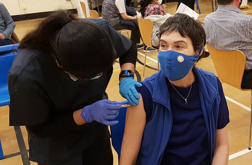 Seated person wearing a mask being injected with a Covid vaccine into their arm by another person leaning over wearing blue disposable gloves.