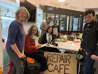 A group of volunteers stand around a table with a cardboard saying &#039;Repair Café&#039; in front of it.